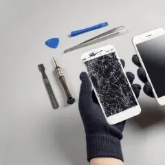 Ręce w rękawiczkach trzymają dwa telefony: jeden z pękniętym ekranem, drugi sprawny. Obok narzędzia do naprawy. Upadek telefonu skutki widoczne.
