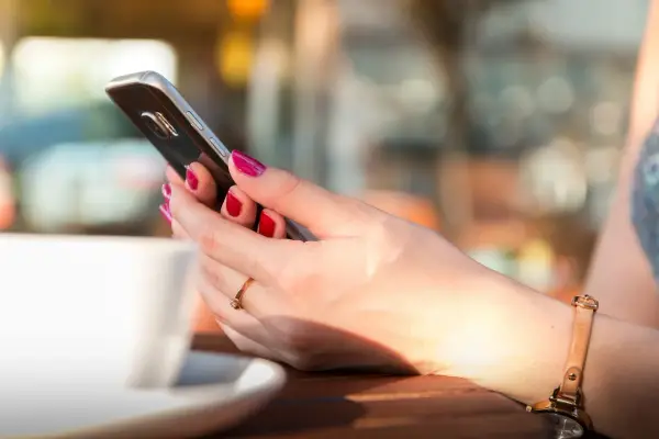 Dłoń z pomalowanymi paznokciami trzyma smartfon, obok biała filiżanka. Długą żywotność baterii w telefonie doceni każda użytkowniczka.